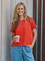 A woman wearing a bright red short-sleeve t-shirt and blue denim shorts with white star prints, holding a takeaway coffee cup against a rustic wooden wall background.