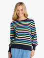 A smiling blonde woman wearing a long-sleeve crewneck sweater with horizontal stripes in navy, green, blue, yellow, pink, and black, paired with light blue jeans, against a plain white background.
