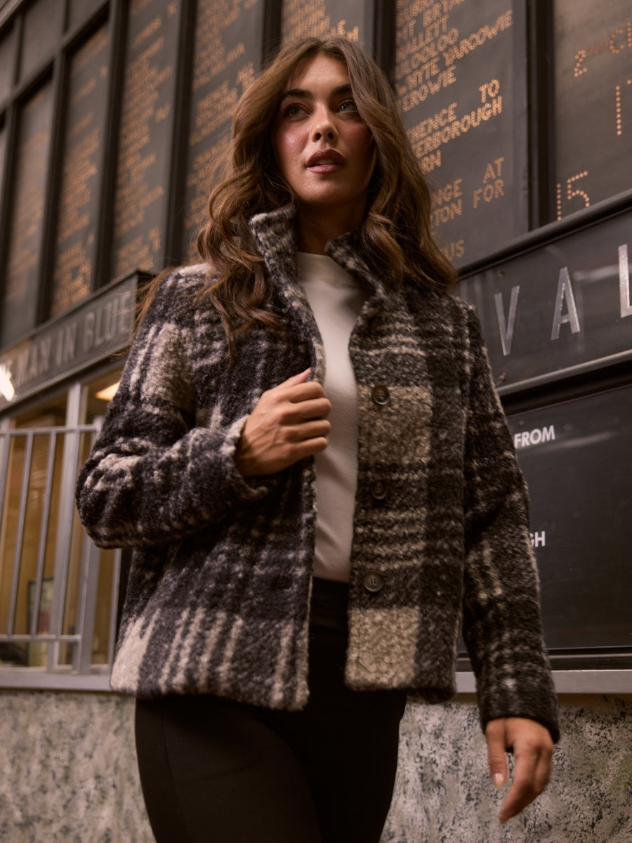Woman wearing a plaid coat standing in front of a building with an electronic sign.