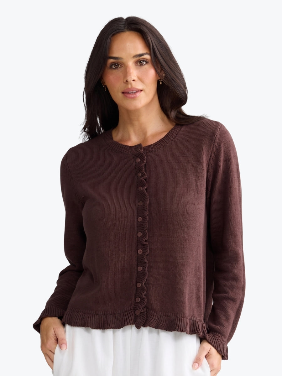 A woman wearing a dark brown ribbed knit cardigan sweater with a scalloped button-front detail, paired with white bottoms, against a plain light gray background.