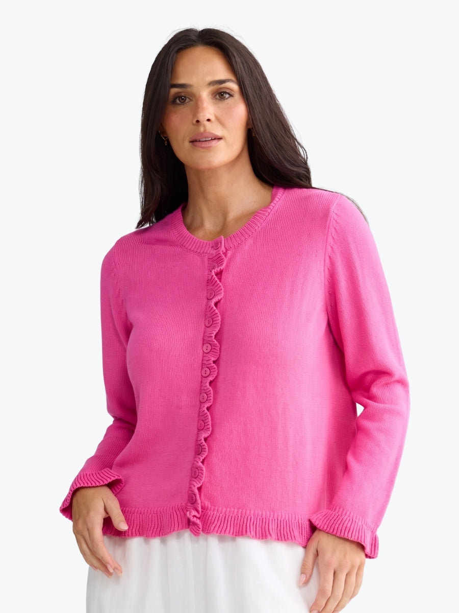 A woman wearing a bright pink knit cardigan sweater with a ruffled button-front detail, shown against a plain light gray studio background.