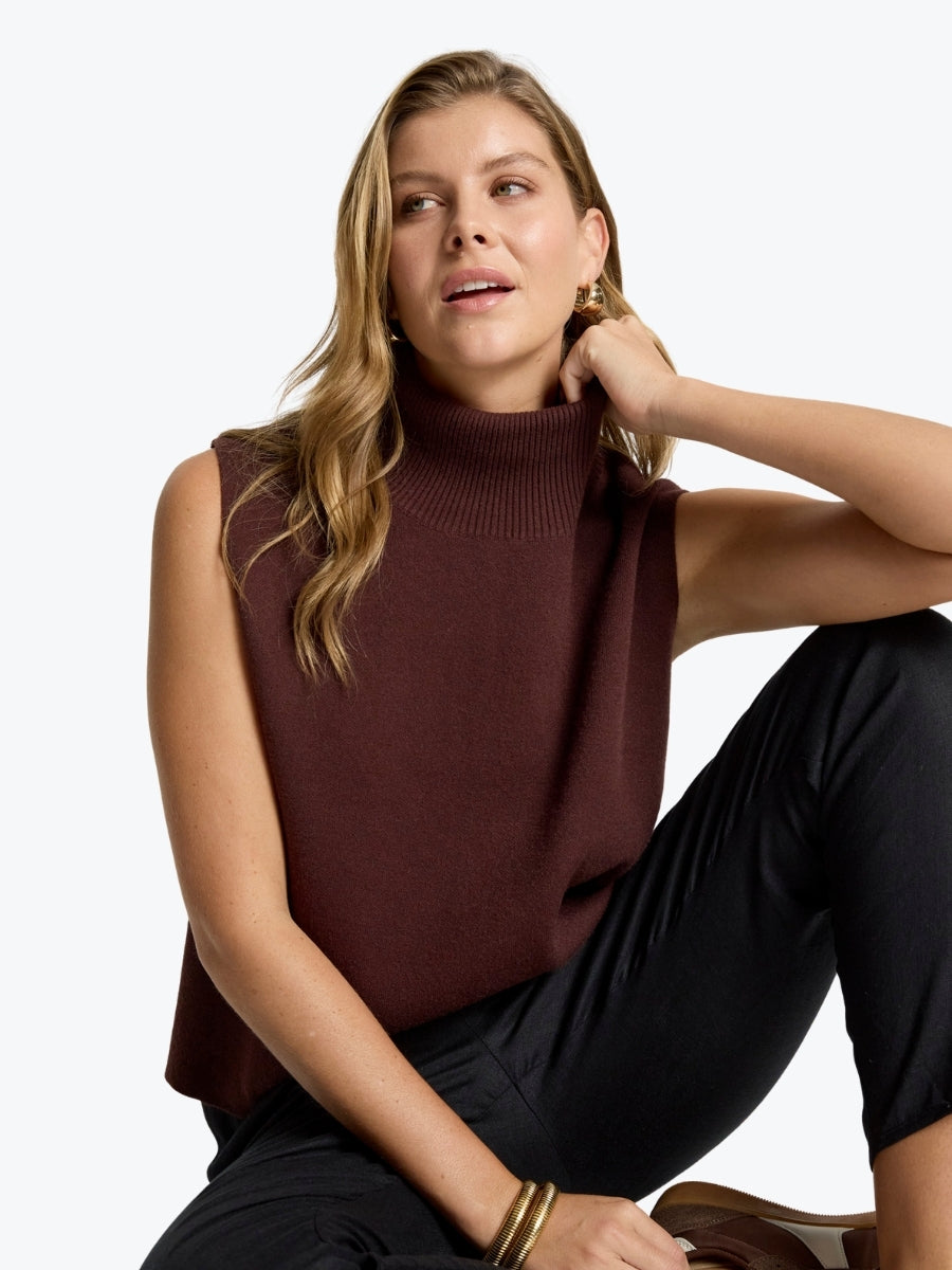 Woman wearing a brown sleeveless sweater and black pants against a white background