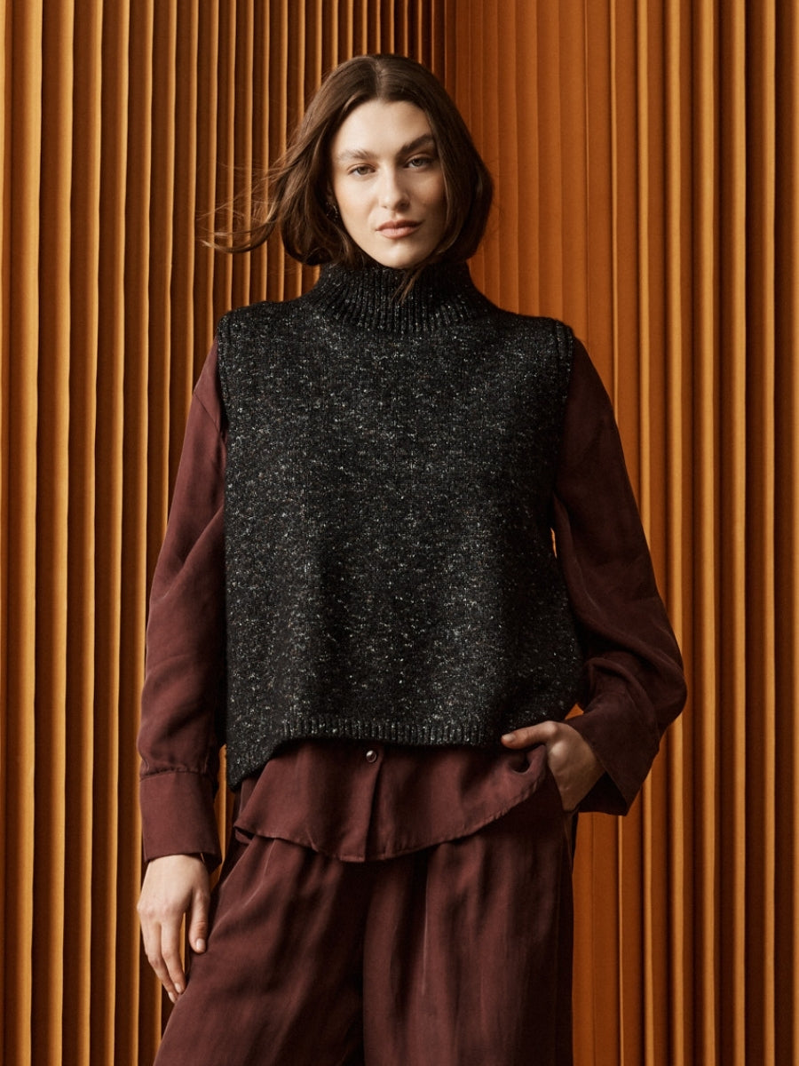 Woman wearing a dark gray knitted vest over a brown shirt against a wooden paneled wall.