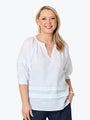 A smiling blonde woman wearing a white three-quarter sleeve blouse with a split neckline and horizontal seam details, paired with dark pants, against a plain white background.