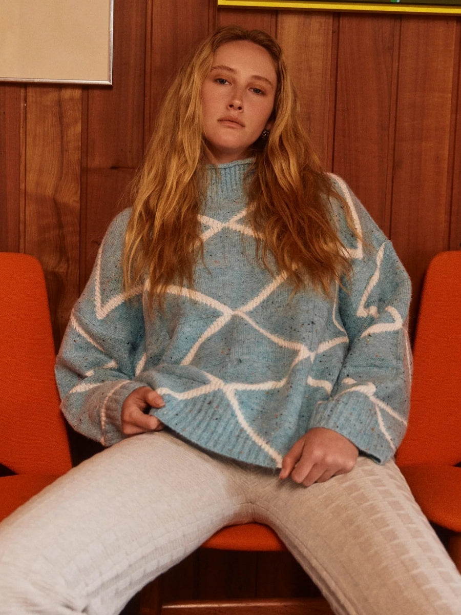 Person wearing a blue and white patterned sweater sitting on an orange chair with a wooden panel background.