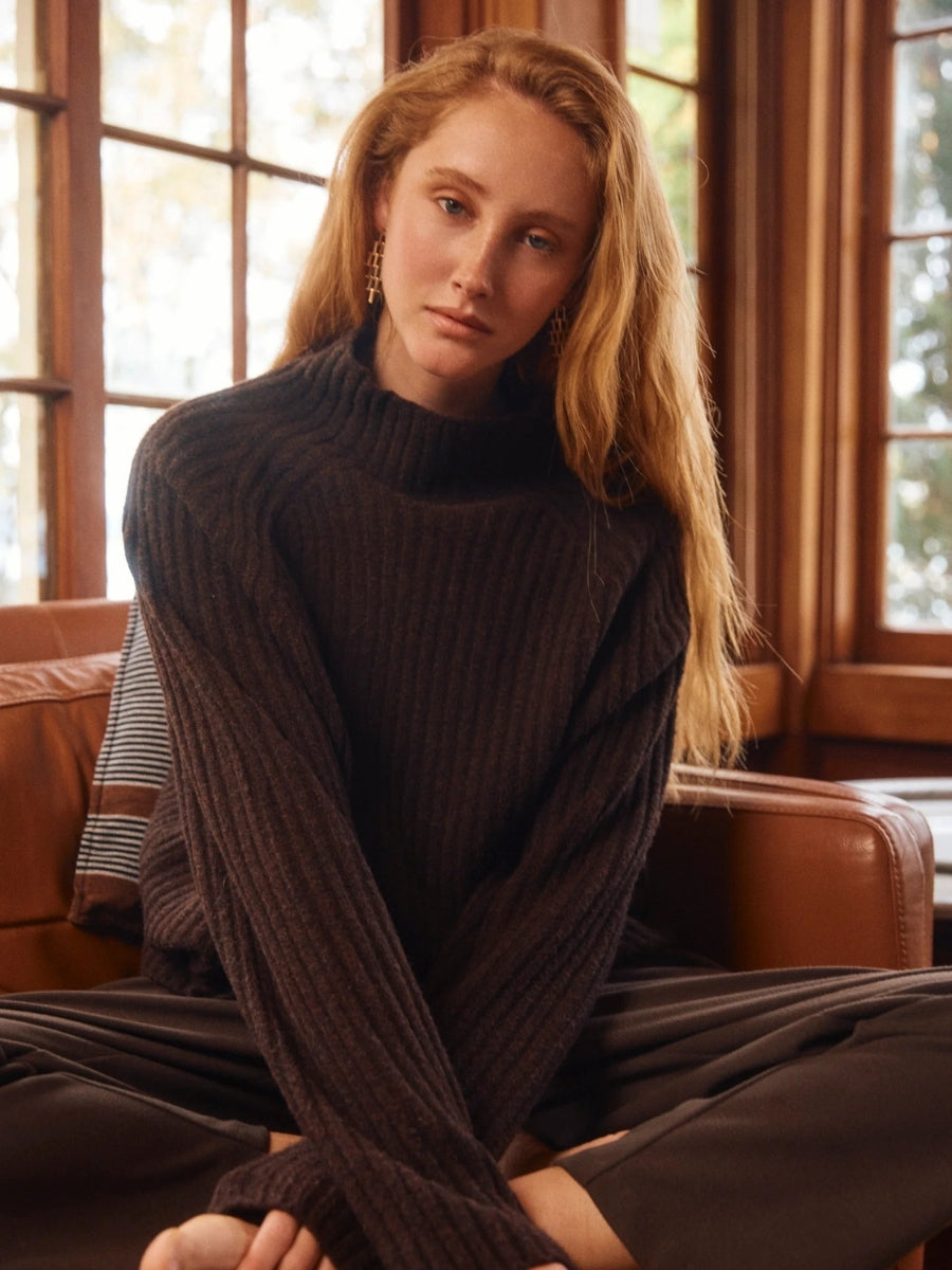 Woman wearing a dark ribbed sweater sitting in a room with large windows.