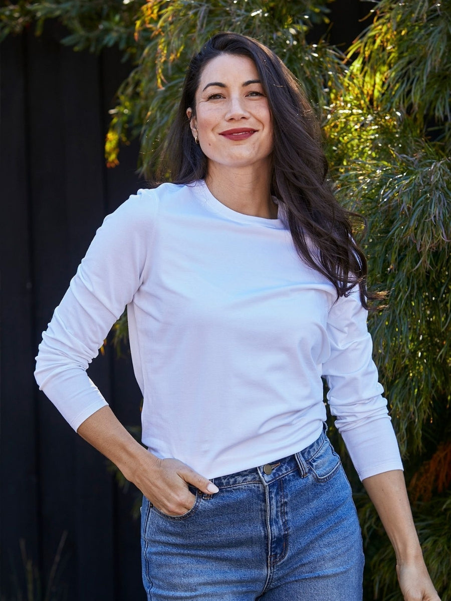 Woman wearing a white long-sleeve shirt and blue jeans standing outdoors with greenery in the background