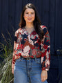 Woman wearing a colorful floral blouse and blue jeans standing against a dark background.