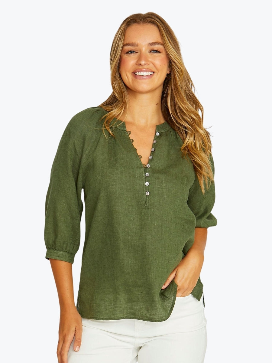 A woman wearing an olive green linen blouse with a buttoned split neckline and white pants, standing against a light gray studio background.
