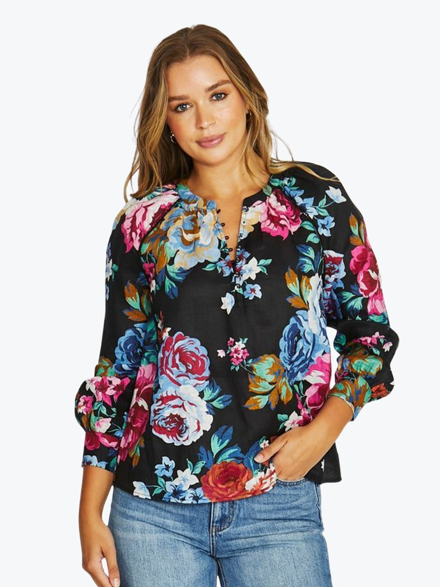 A woman wearing a black long-sleeve blouse with a vibrant multicolor floral print, paired with blue jeans, against a plain light gray background.