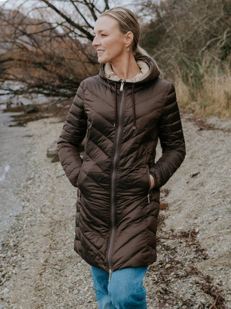 Woman wearing a brown puffer jacket standing outdoors near a body of water.