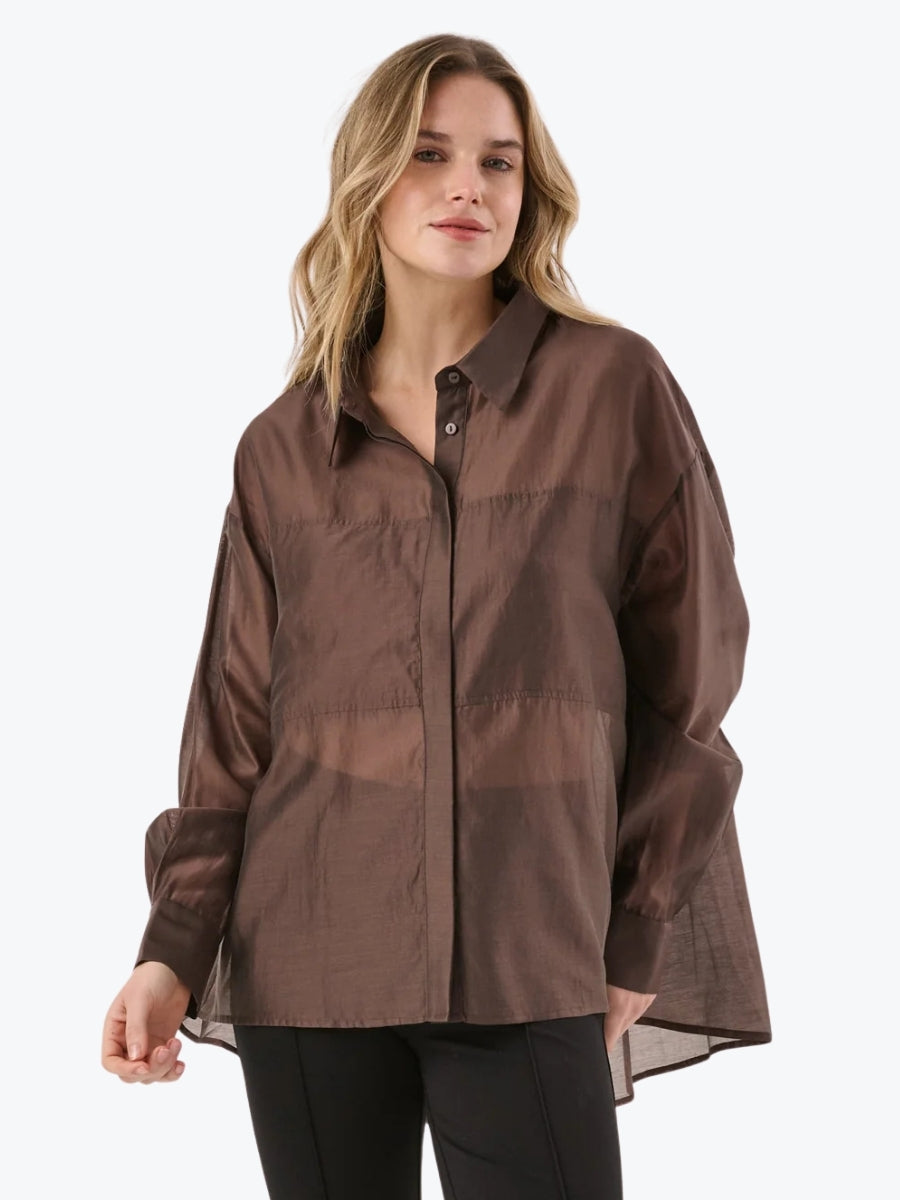 A woman wearing a brown button-up shirt with sheer mesh panel details, paired with black pants, against a plain white background.