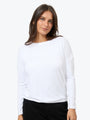 A woman wearing a white long-sleeve boat-neck top and black pants, photographed against a clean light gray studio background.