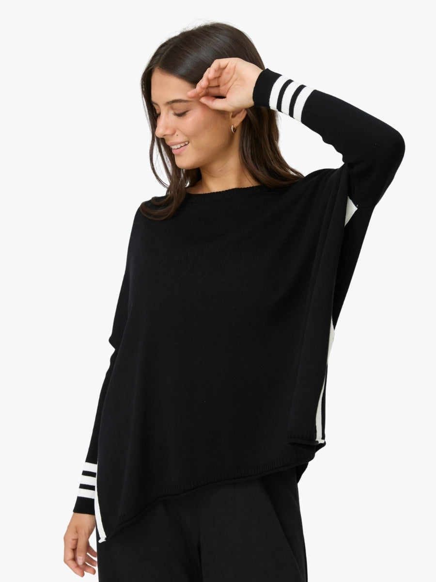 A woman wearing a black long-sleeve knit top with white stripe accents on the cuffs and side seams, photographed against a plain light gray studio background.