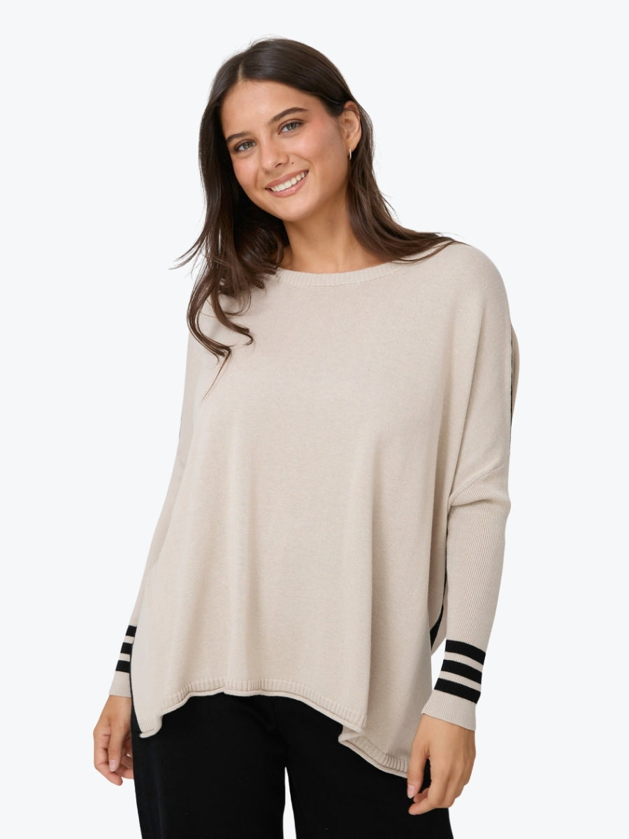 A woman wearing a light beige long-sleeve knit top with black and white striped cuffs, paired with black pants, against a plain white background.