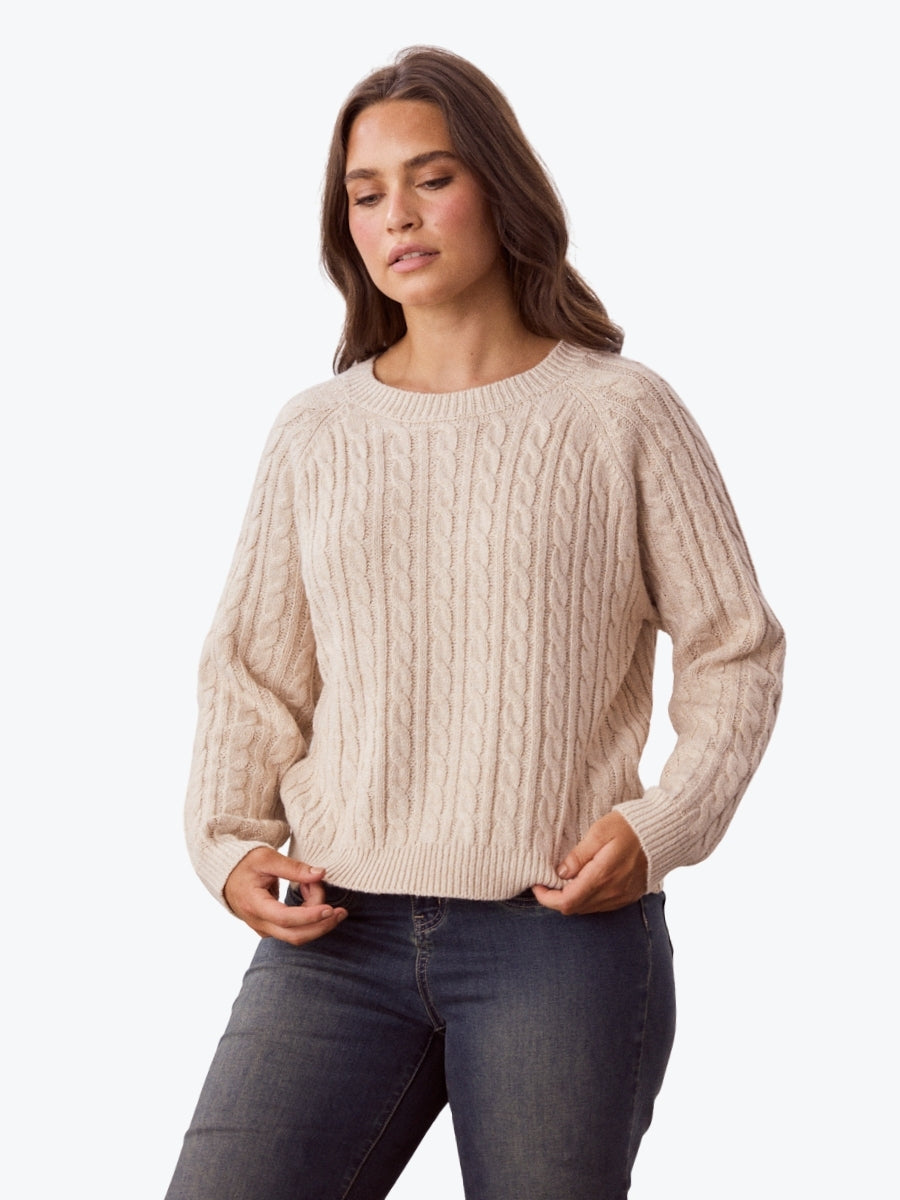 Woman wearing a beige cable knit sweater and dark jeans on a white background