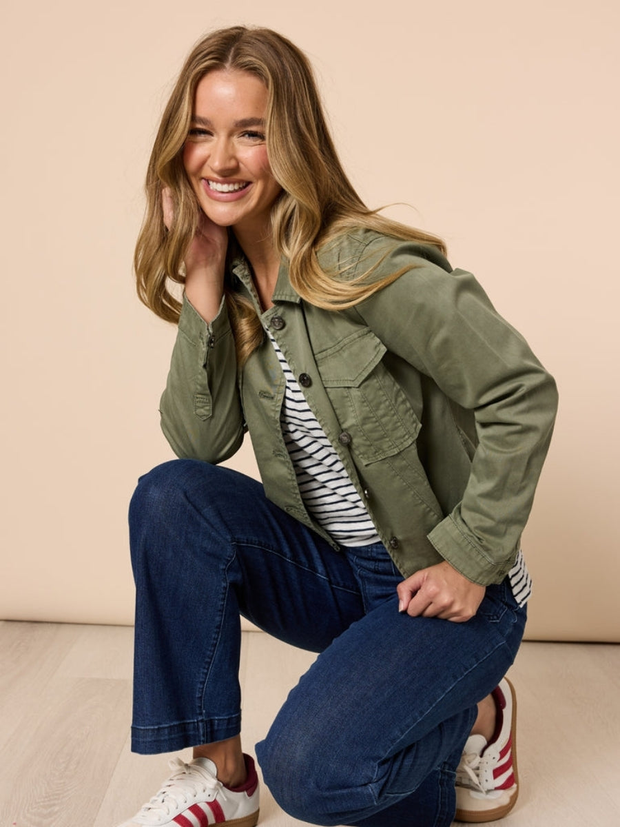 Woman wearing a khaki jacket, striped shirt, and blue jeans on a beige background